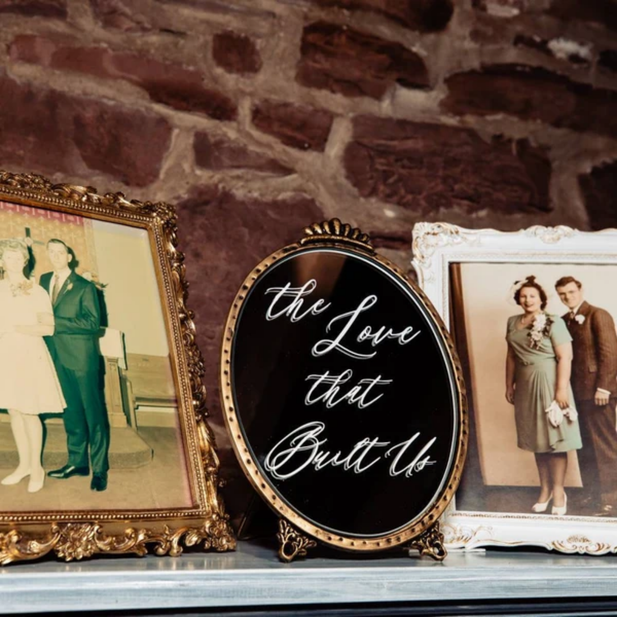 An oval black sign with gold trim and white script reading 'the Love that Built Us' displayed on a mantel between two vintage framed photographs against a rustic stone wall.