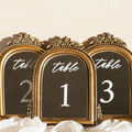 Three ornate gold-framed table number signs with black backgrounds and white script text reading 'table' above the numbers 2, 1, and 3, styled with white fabric at the bottom against a light beige background.
