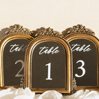 Three ornate gold-framed table number signs with black backgrounds and white script text reading 'table' above the numbers 2, 1, and 3, styled with white fabric at the bottom against a light beige background.