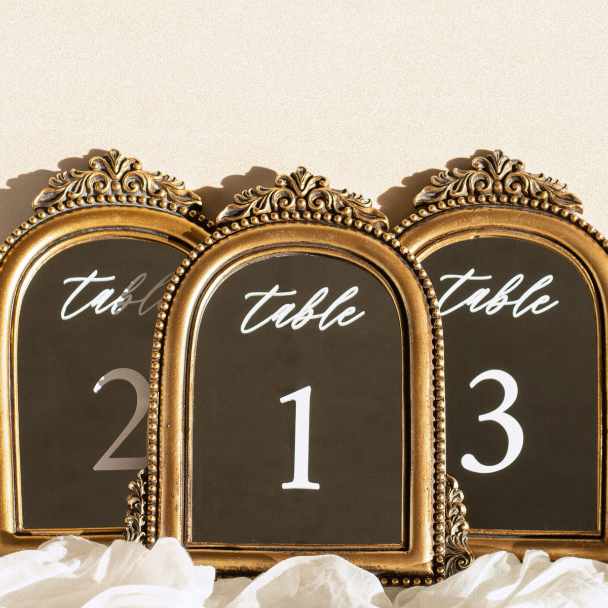 Three ornate gold-framed table number signs with black backgrounds and white script text reading 'table' above the numbers 2, 1, and 3, styled with white fabric at the bottom against a light beige background.