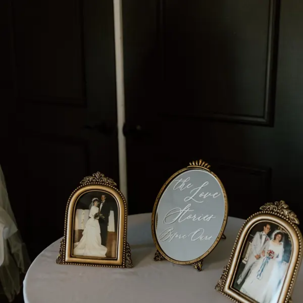 Three ornate gold-framed photo frames displayed on a white tablecloth-covered table against a dark paneled wall. The center frame is oval and reads 'the Love Stories Begin Our' in cursive, flanked by two vertical portrait frames containing wedding photographs.