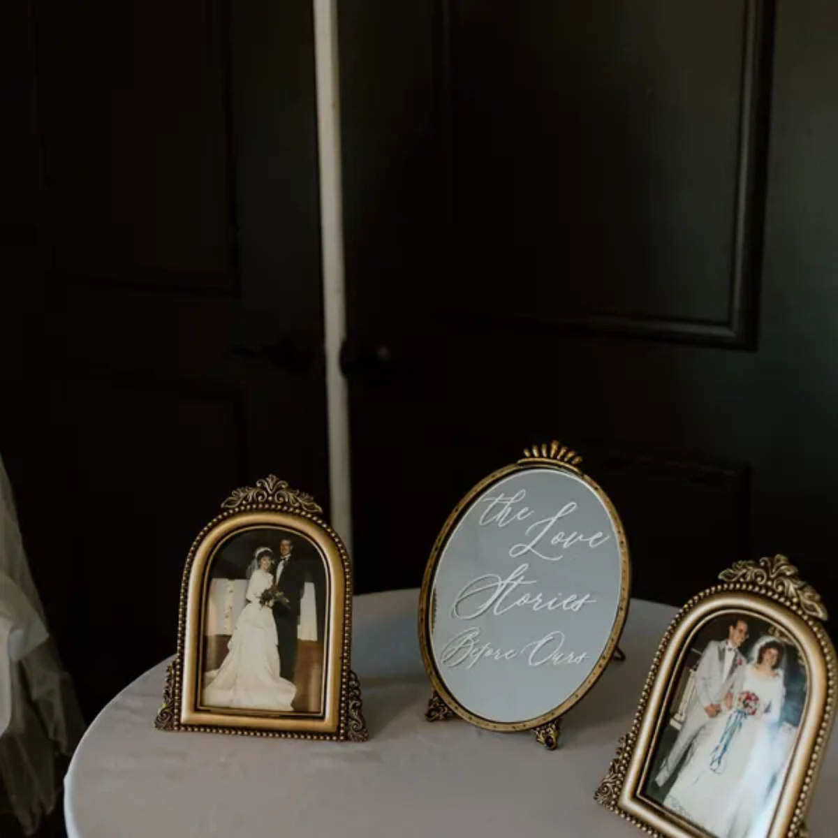 Three ornate gold-framed photo frames displayed on a white tablecloth-covered table against a dark paneled wall. The center frame is oval and reads 'the Love Stories Begin Our' in cursive, flanked by two vertical portrait frames containing wedding photographs.