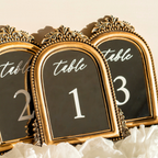 Three ornate gold table number signs with arched tops and black faces displaying white text reading 'table' and numbers 1, 2, and 3, arranged on white ruffled fabric against a warm beige background.