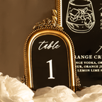 An ornate gold-framed table number sign displaying 'table 1' in white lettering, styled on white ruffled fabric against a dark brown background with a matching sign partially visible behind it.