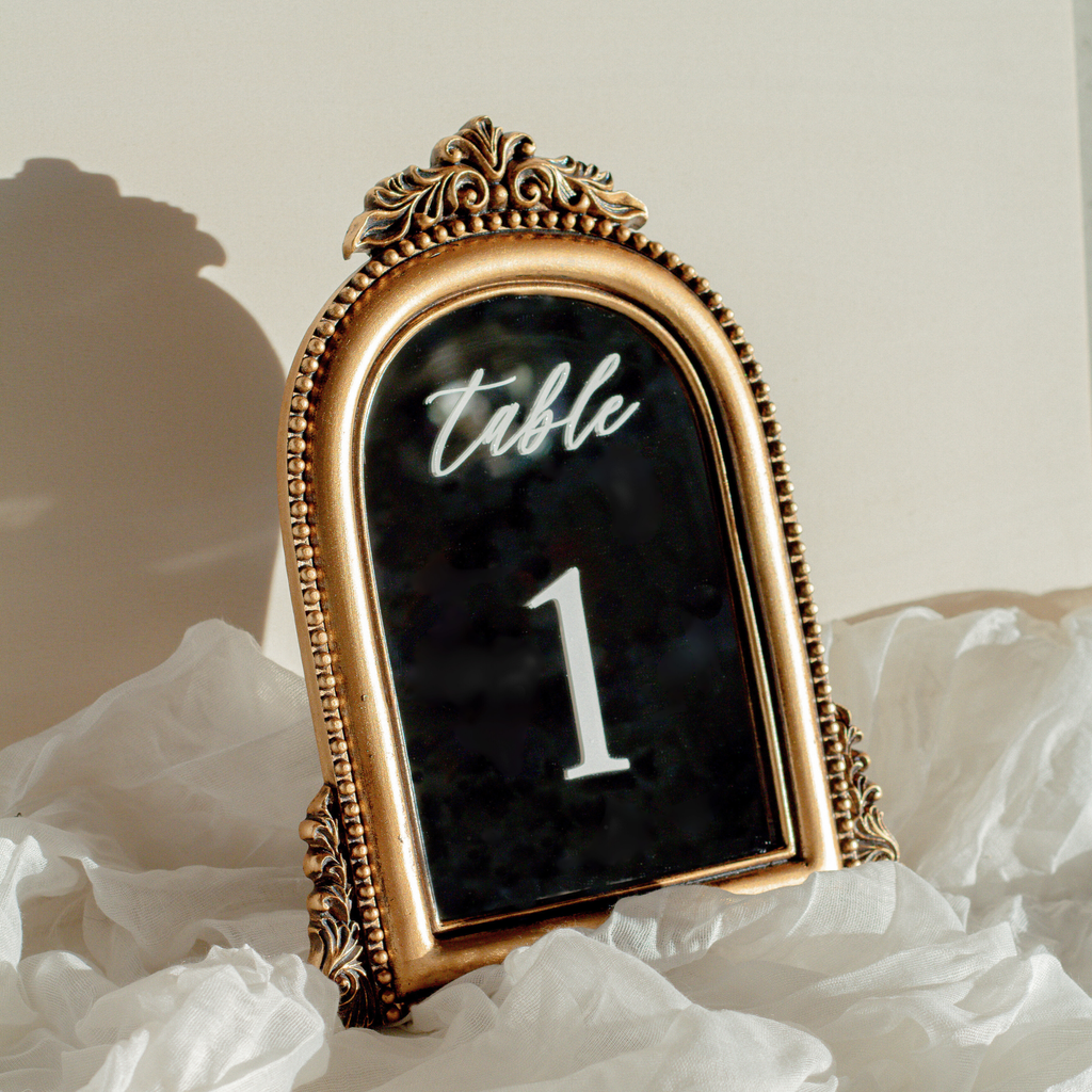 An ornate gold table number frame displaying 'Table 1' in white script and numerals on a black background, styled on white ruffled fabric.