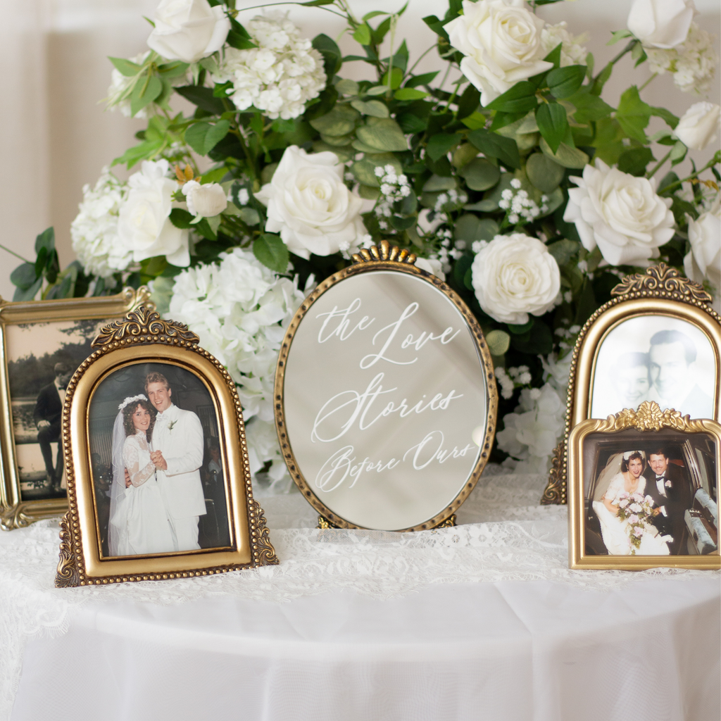 A styled wedding display featuring three ornate gold picture frames and a central oval mirror with the phrase 'the Love Stories Before Us,' set against a large white rose and green foliage arrangement on a white tablecloth.