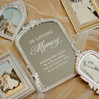 An ornate silver arched photo frame with a gray insert that reads 'IN LOVING Memory of those who cannot be with us today, but are forever with us in our hearts,' styled on a wooden surface with tulle and other decorative frames.