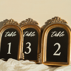 Three ornate gold-framed table number signs with black backgrounds and white lettering reading 'table' above the numbers 1, 3, and 2, styled on white fabric against a warm beige wall.
