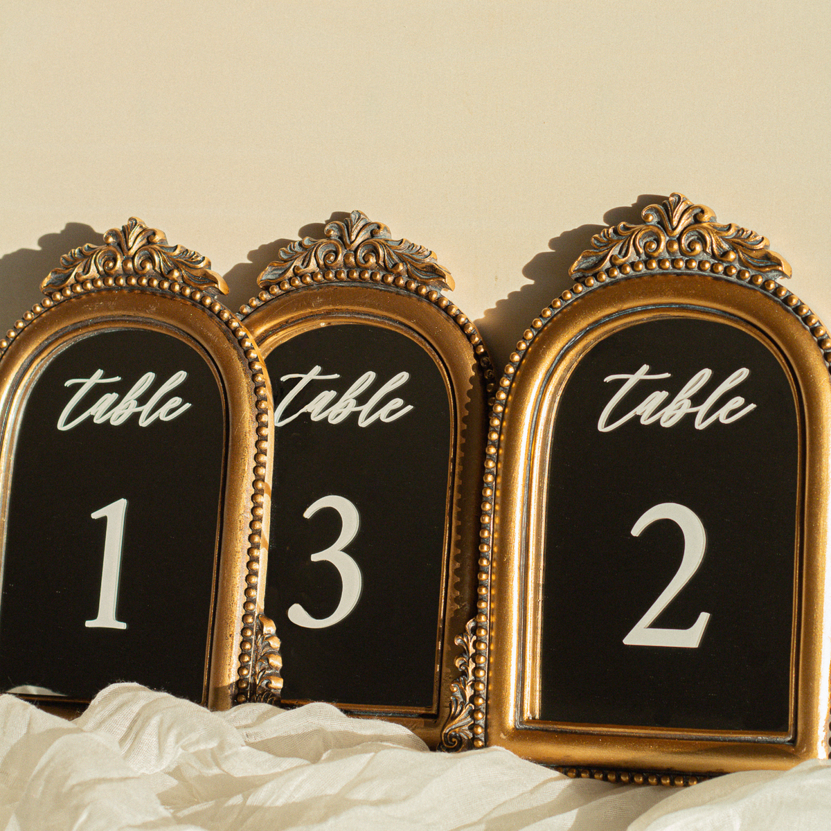Three ornate gold-framed table number signs with black backgrounds and white lettering reading 'table' above the numbers 1, 3, and 2, styled on white fabric against a warm beige wall.