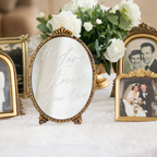 An oval tabletop mirror with a gold-toned frame and the phrase 'the Love Stories Before Our' in cursive script, styled on a white tablecloth with white roses and other framed wedding photos in the background.