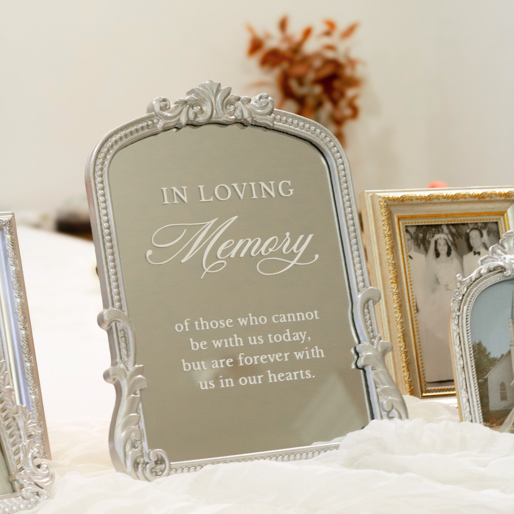 An ornate silver mirror with a memorial message that reads 'IN LOVING Memory of those who cannot be with us today, but are forever with us in our hearts.' displayed on a white draped surface next to other picture frames.