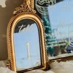 An ornate gold-framed table number sign displaying 'table 1' in elegant script and numerals, styled on white textured paper with a blurred cocktail menu in the background.