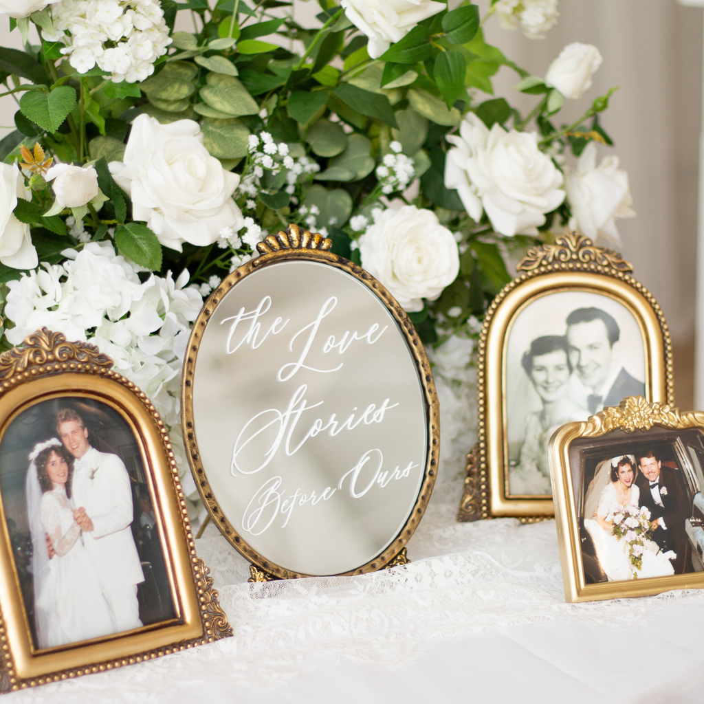 A styled wedding display featuring four ornate gold picture frames with vintage photographs arranged around a central oval mirror that reads 'the Love Stories Before Our'. The setup is decorated with lush white roses and green foliage on a white lace-covered surface.