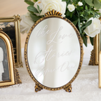 An oval tabletop mirror with an ornate gold frame and matching gold stand, featuring the phrase 'the Love Stories Before Our' in cursive script across the reflective surface. The mirror is styled on a white lace surface alongside other gold picture frames and white roses.