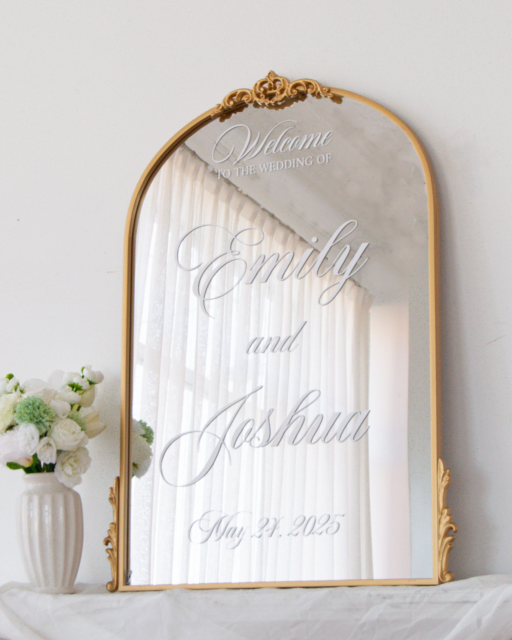 An arched gold-framed mirror with ornate corner details, reflecting white curtains and personalized with the text 'Welcome to the wedding of Emily and Joshua May 21, 2025' in elegant silver script. The mirror is styled on a white draped surface next to a white ceramic vase with white and green flowers against a white wall.