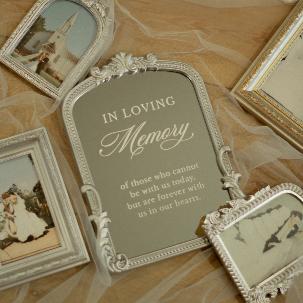 An ornate silver memorial picture frame with a gray insert displaying the text 'IN LOVING Memory of those who cannot be with us today, but are forever with us in our hearts.' The frame is styled on a light wood surface with tulle fabric and other decorative frames around it.