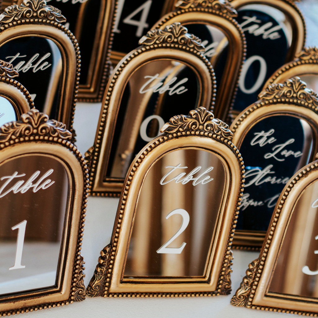 A close-up of ornate gold table number frames arranged on a table, with visible numbers 1 through 3 and script text reading 'table' above each number.
