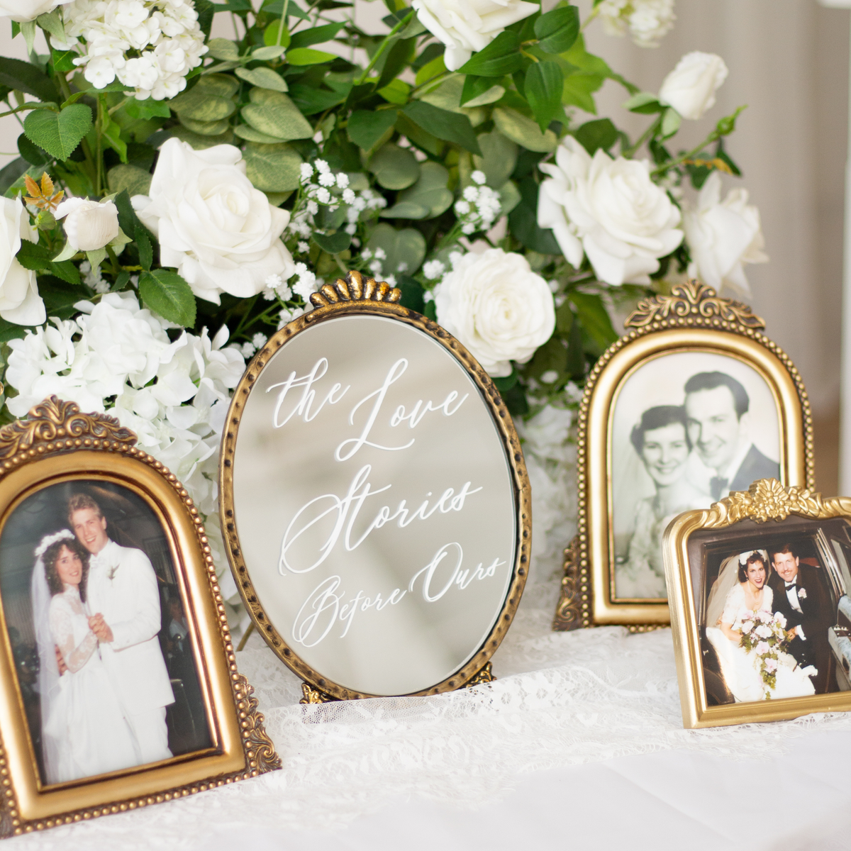 A styled wedding display featuring four ornate gold picture frames with vintage photographs arranged around a central oval mirror that reads 'the Love Stories Before Our'. The setup is decorated with lush white roses and green foliage on a white lace-covered surface.