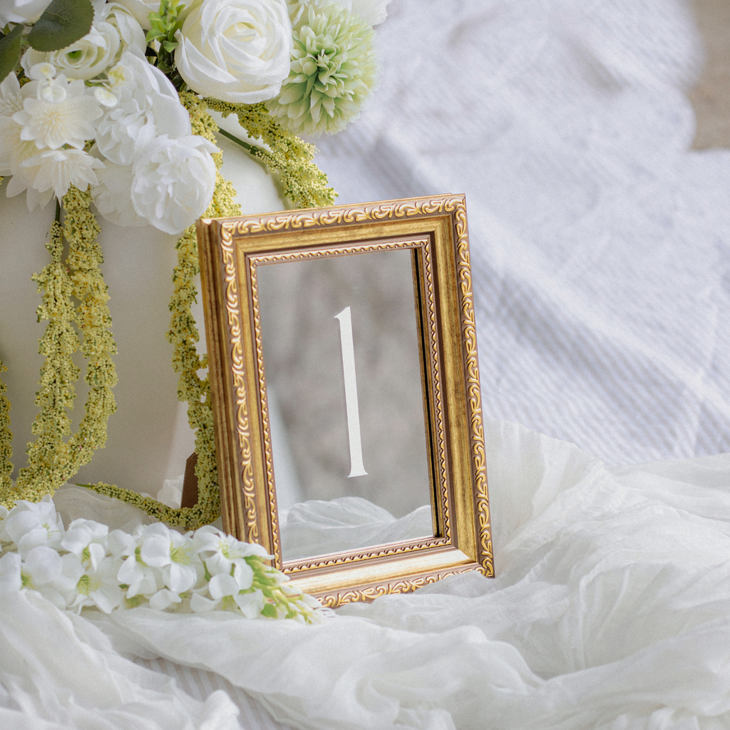An ornate gold table number frame displaying the number 1, styled with white flowers and draped white fabric.