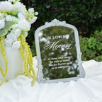 An ornate silver-framed mirror with a reflective glass surface displaying the words 'IN LOVING Memory' and a memorial quote, styled on white draped fabric next to a white floral arrangement in an outdoor garden setting.