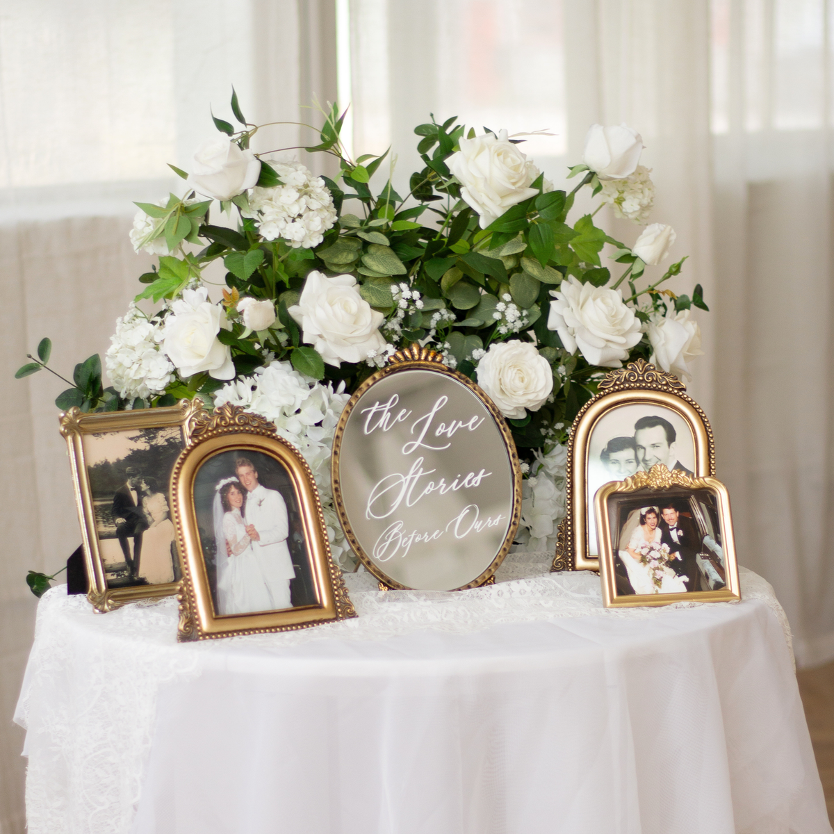 A styled wedding-themed display on a white draped table featuring a large white rose and green foliage arrangement, several ornate gold-framed photo frames with wedding photos, and a central oval mirror that reads 'the Love Stories Before Our'.