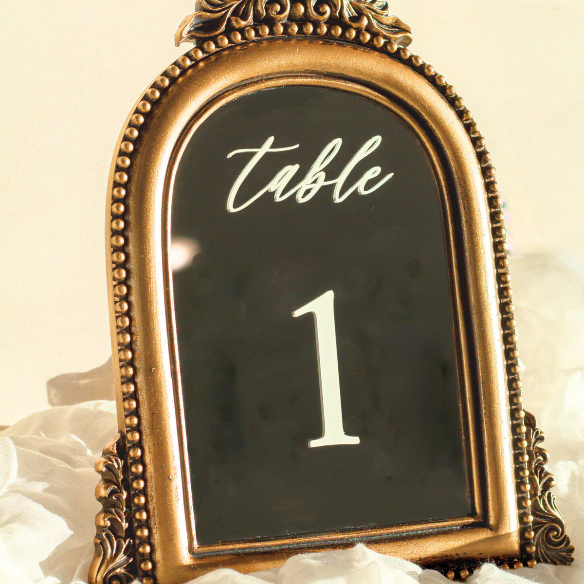An ornate gold-framed table number sign displaying 'table 1' in white lettering, set against a soft cream background with white fabric accents.
