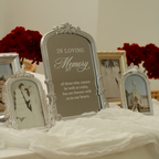 A silver arched memorial photo frame with engraved text reading 'IN LOVING Memory' is displayed on a white draped tablecloth, flanked by smaller matching photo frames and red rose arrangements in the background.