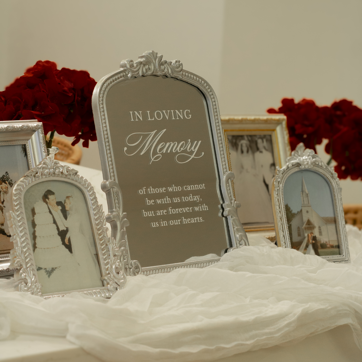 A silver arched memorial photo frame with engraved text reading 'IN LOVING Memory' is displayed on a white draped tablecloth, flanked by smaller matching photo frames and red rose arrangements in the background.