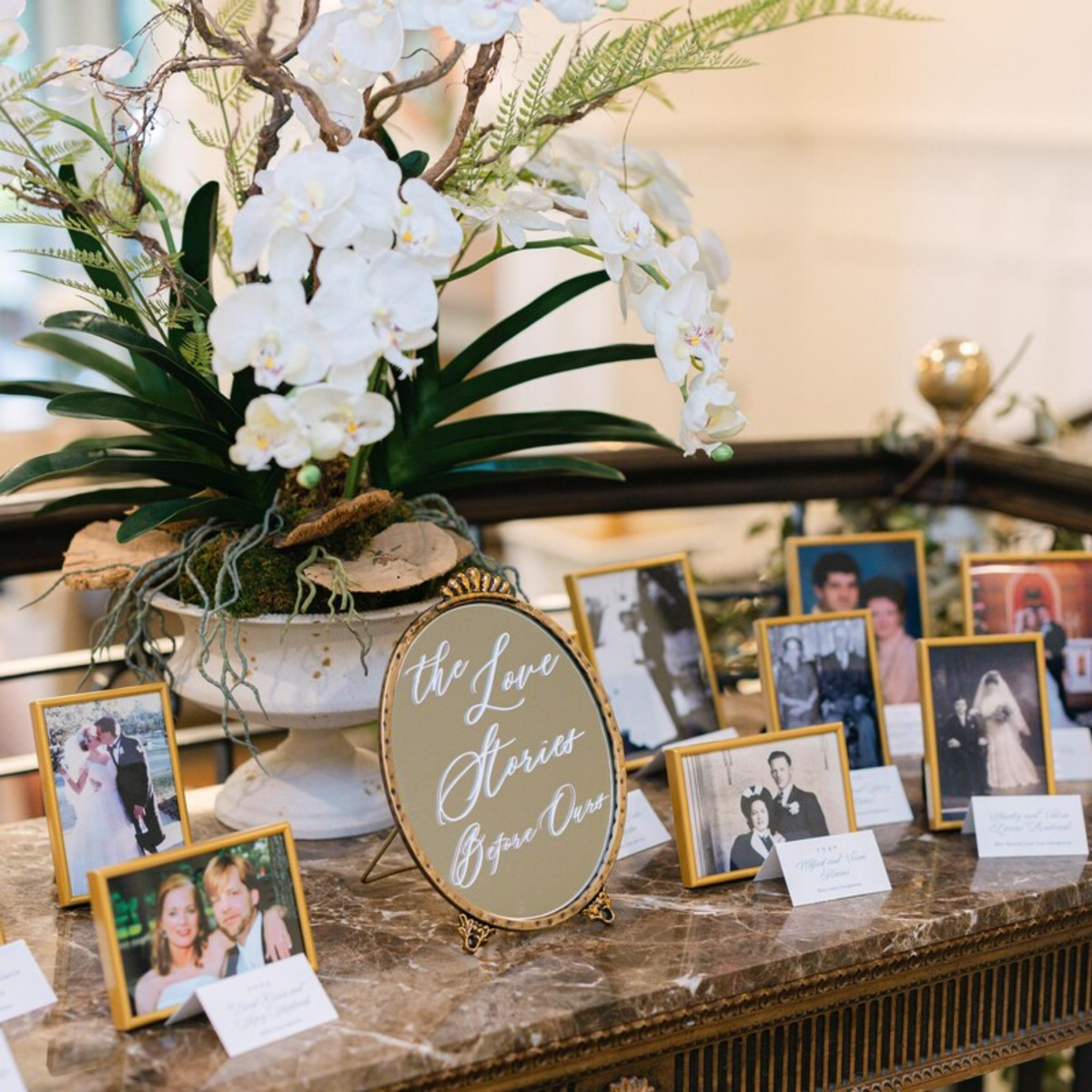 A wedding reception display on a marble table featuring a large white orchid arrangement in a white urn, a round gold-framed sign reading 'the Love Flowers Bring One,' and multiple small framed photos with handwritten name cards.