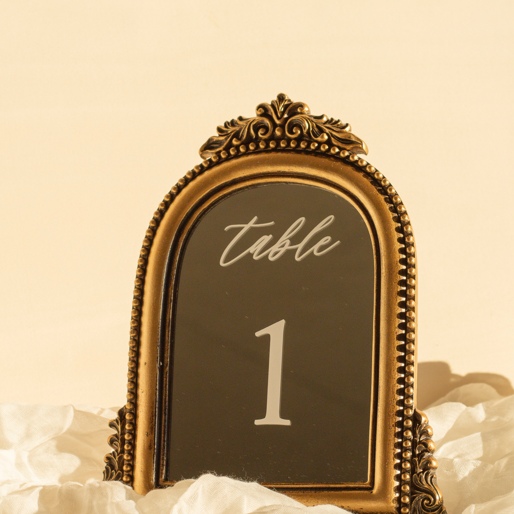 An ornate antique-style gold table number plaque displaying 'table 1' in white lettering on a dark background, styled on white tissue paper against a warm beige backdrop.