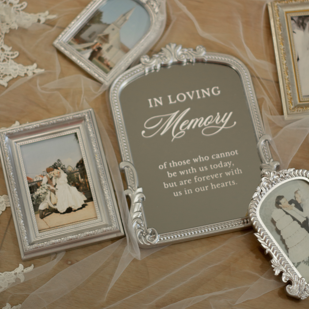 A decorative memorial photo frame with a gray center panel reading 'IN LOVING Memory of those who cannot be with us today, but are forever with us in our hearts,' surrounded by ornate silver frames containing wedding and family photographs on a beige fabric backdrop with lace and tulle.