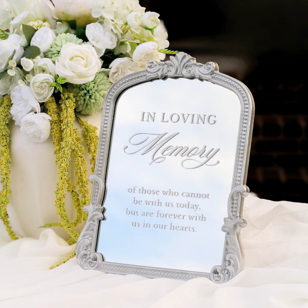 An ornate silver memorial photo frame with a printed light blue insert reading 'In Loving Memory' and a floral arrangement in the background.