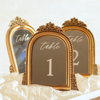 Three ornate gold table number signs with arched tops and decorative frames, displaying table numbers 1, 2, and 3, arranged on white fabric.
