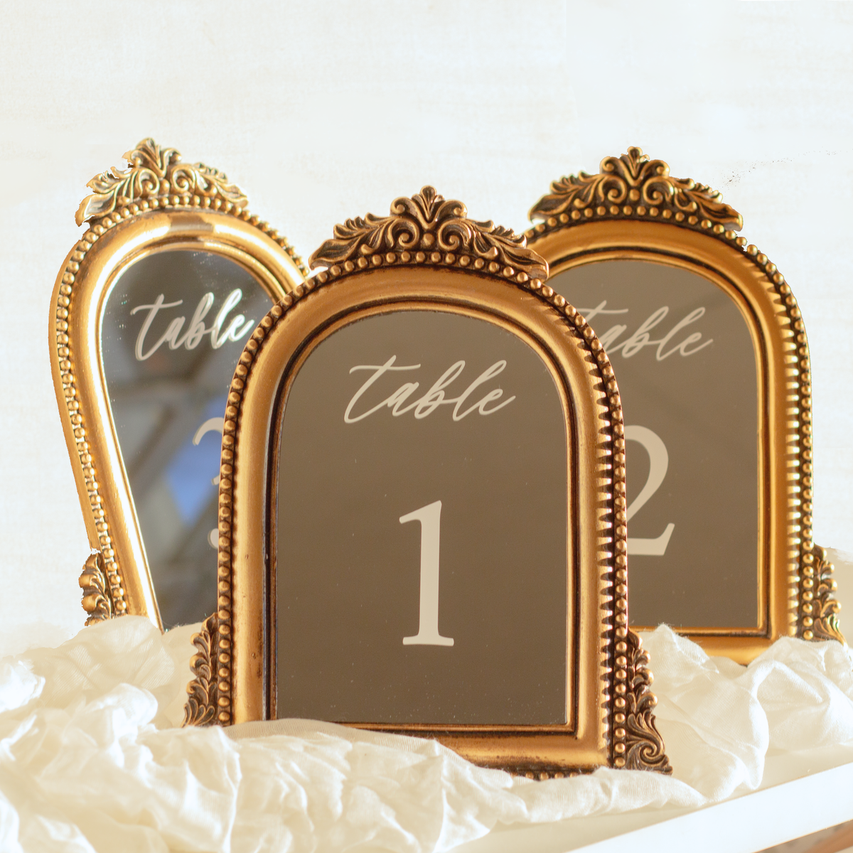 Three ornate gold table number signs with arched tops and decorative frames, displaying table numbers 1, 2, and 3, arranged on white fabric.